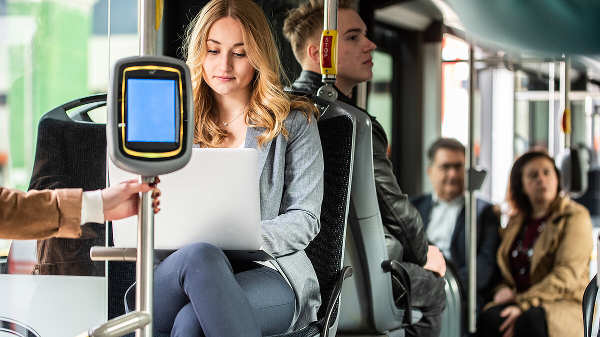 Una ragazza seduta in autobus lavora con il laptop. Sullo sfondo si vedono altri passeggeri.