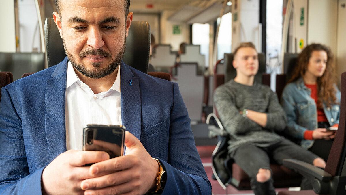 Un passeggero seduto in treno controlla il cellulare. Sullo sfondo a destra si vede una coppia di passeggeri seduti.
