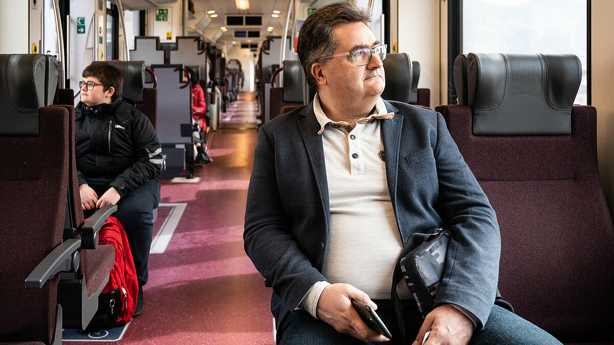 Un passeggero seduto in treno con in mano un cellulare guarda fuori dal finestrino. Si vede un altro passeggero dietro di lui sulla sinistra.