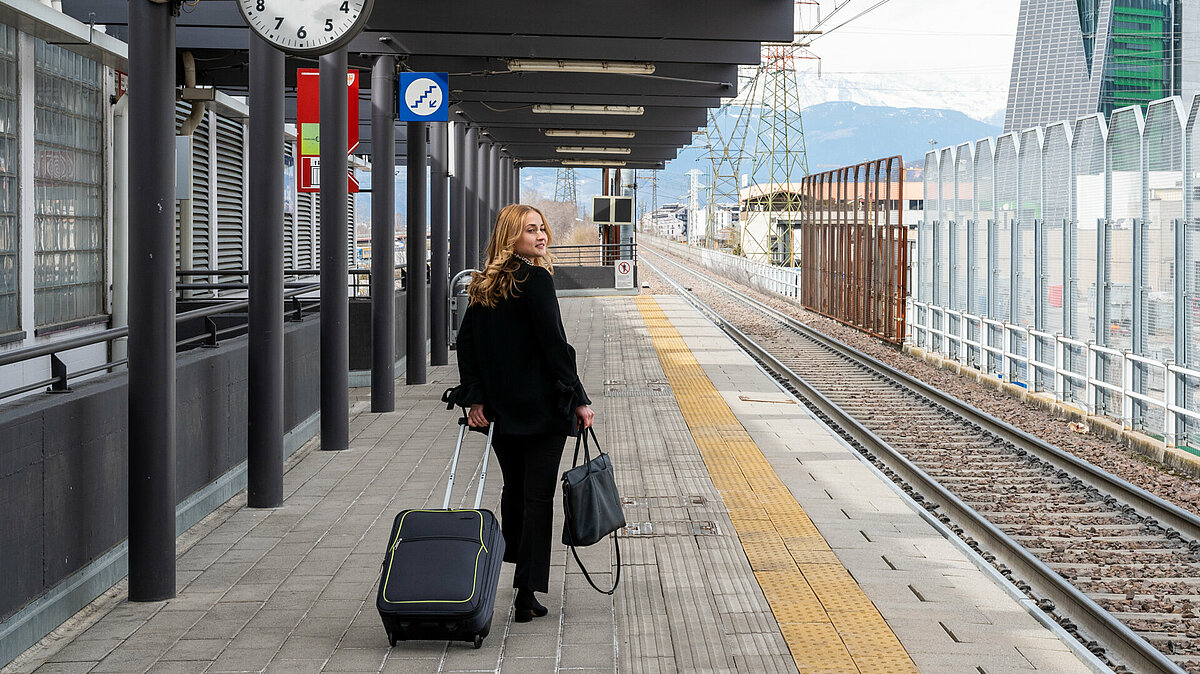 Una ragazza con borsa e valigia cammina lungo la banchina della stazione. Guarda verso i binari.